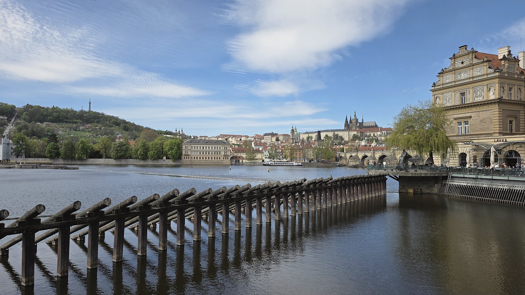 View north from Smetanovo nábřeží: wooden ice-breakers in the foreground, Karlův most spanning to Malá Strana, Pražský hrad and the spires of St. Vitus rising on the right, the corner of the Bedřich Smetana Museum at the far right.